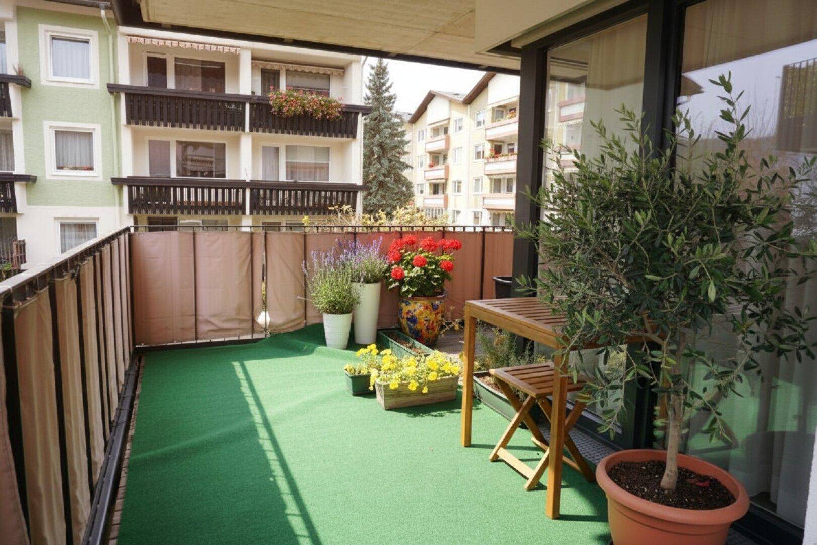 Langer Balkon mit Kunstrasen, Holztisch und Stühlen, geschmückt mit bunten Topfpflanzen.