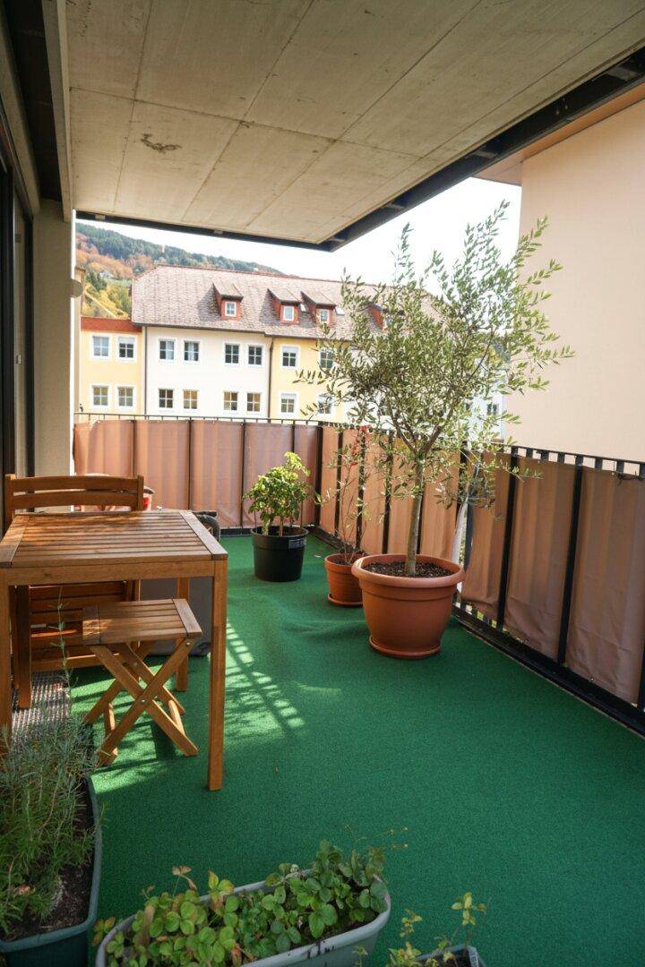 Überdachter Balkon mit Kunstrasen, Holztisch und einem großen Olivenbaum, bietet Bergblick.