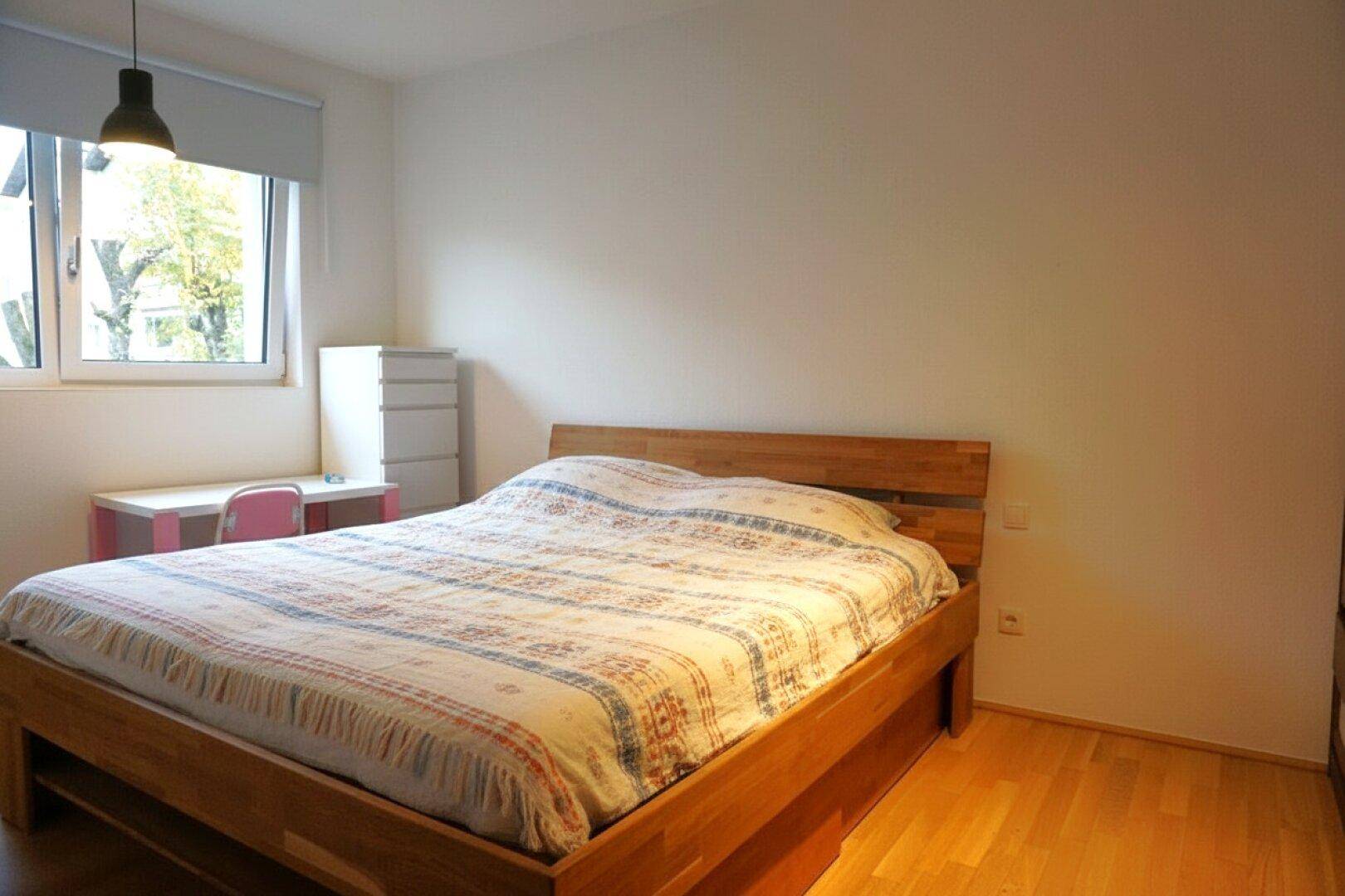 Helles Schlafzimmer mit Holzbett, Schreibtisch am Fenster und Parkettboden, ideal für Ruhe.