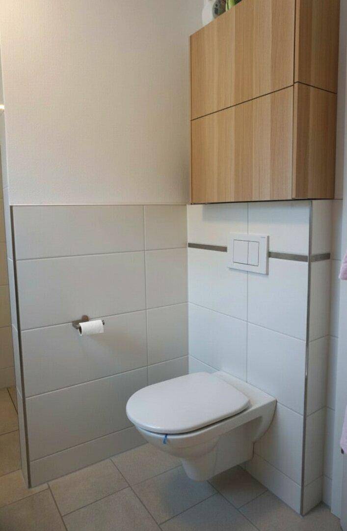 Wandhängendes WC mit Holzschrank darüber und hellen Fliesen, funktional und sauber.