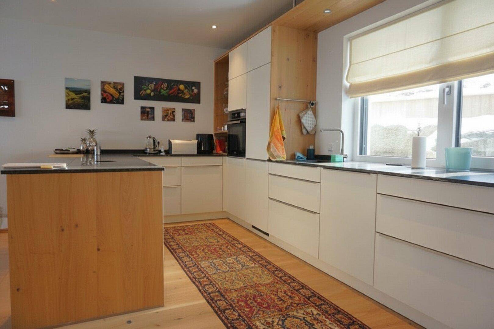Moderne Küche mit Holzelementen, weißen Fronten, Kücheninsel und Fenster mit Blick ins Grüne.