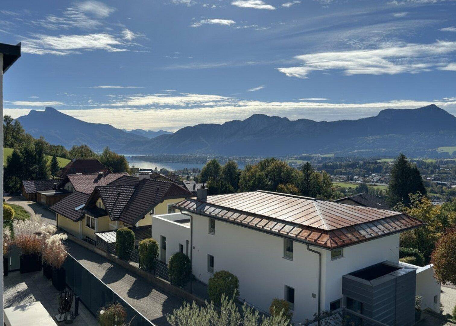 Panoramablick auf die umliegende Landschaft mit Bergen, See und Wohnbebauung unter blauem Himmel.