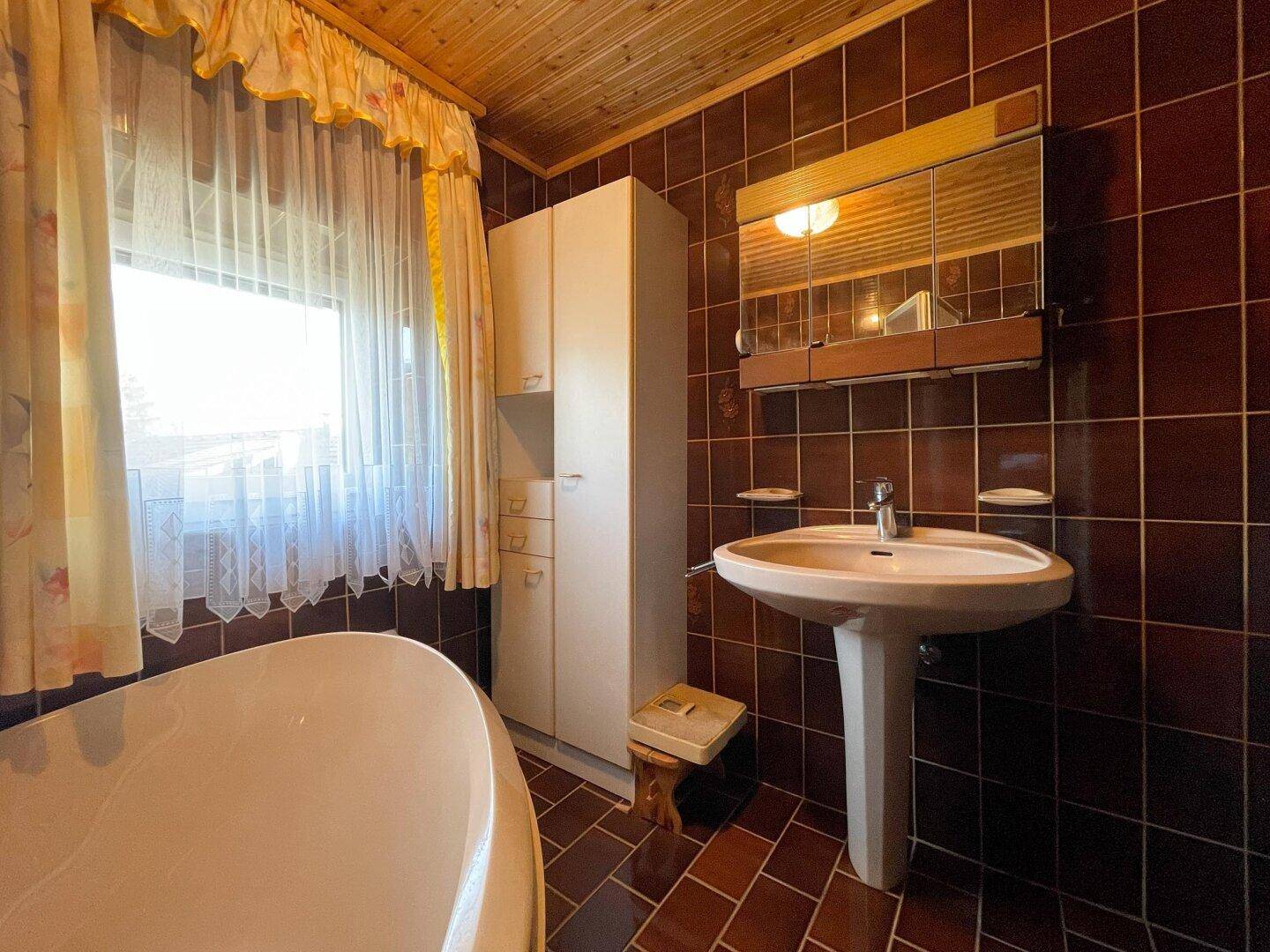Badezimmer mit braunen Fliesen, Waschbecken und Spiegelschrank, beleuchtet durch ein Fenster.