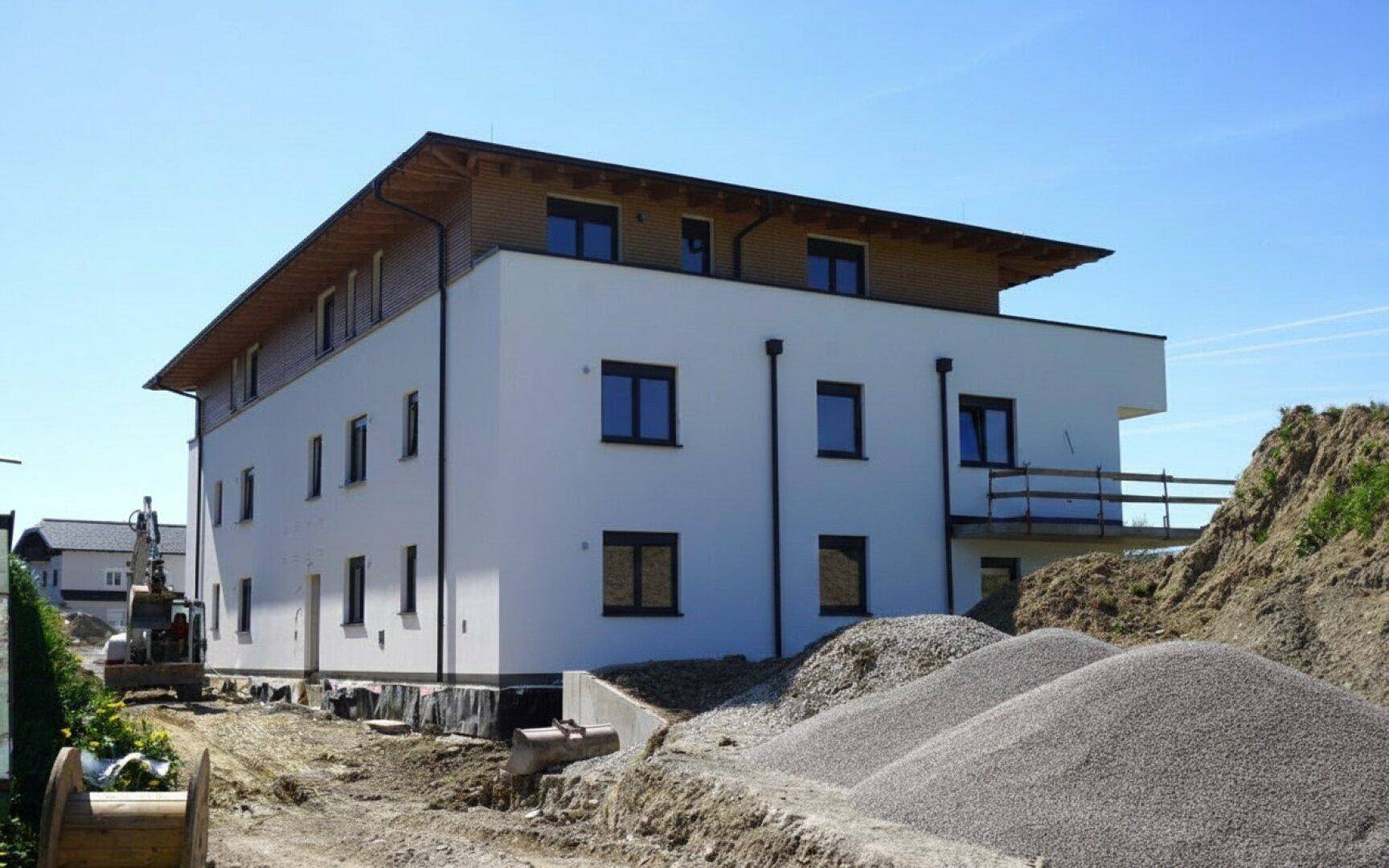 Rückseite des Neubaus mit Bauarbeiten und Materialhaufen, umgeben von einer ländlichen Landschaft mit Bergen.