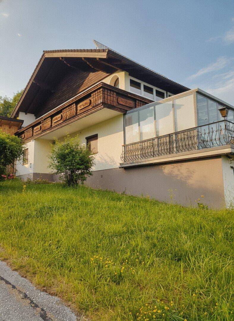 Seitenansicht des Hauses mit verglastem Wintergarten und traditionellem Holzbalkon bei Sonnenschein.