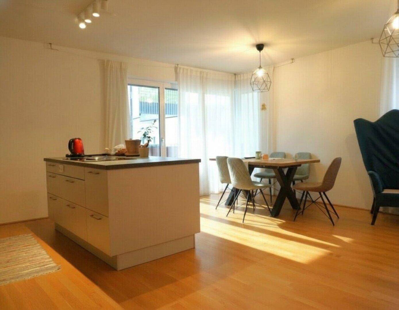 Offener Wohn- und Essbereich mit Kücheninsel, Esstisch und Zugang zu einem Balkon, hell beleuchtet.