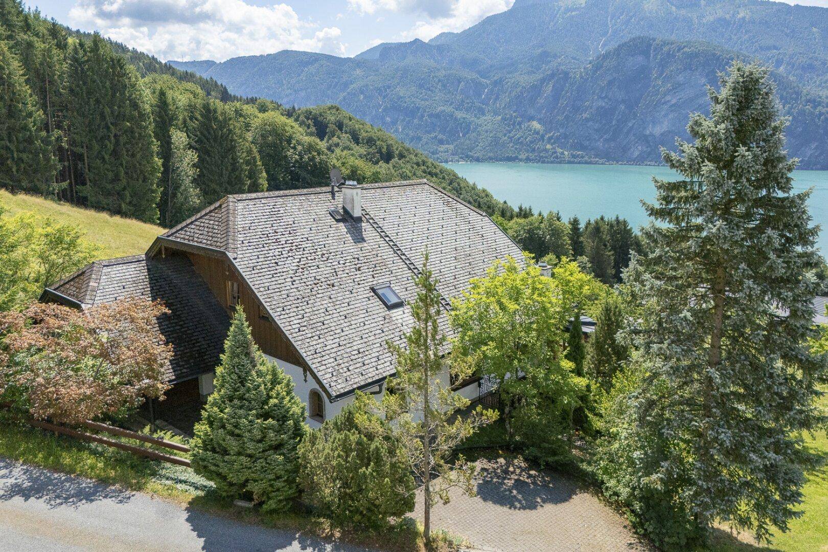 Luftaufnahme eines Hauses mit traditionellem Dach, umgeben von Bäumen und Bergen mit Seeblick.