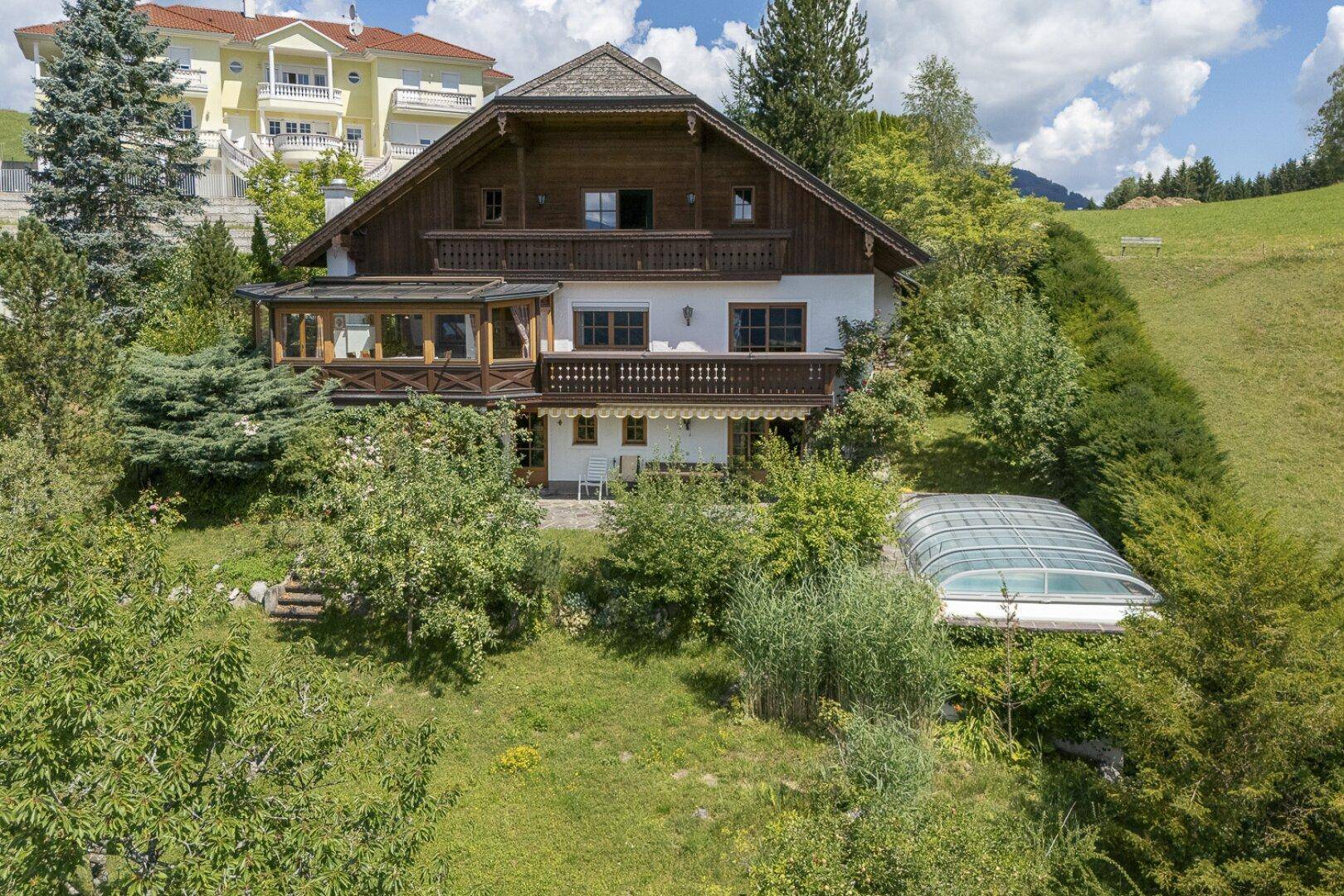 Traditionelles Haus mit Holzbalkonen und einem Wintergarten, umgeben von einem grünen Garten.