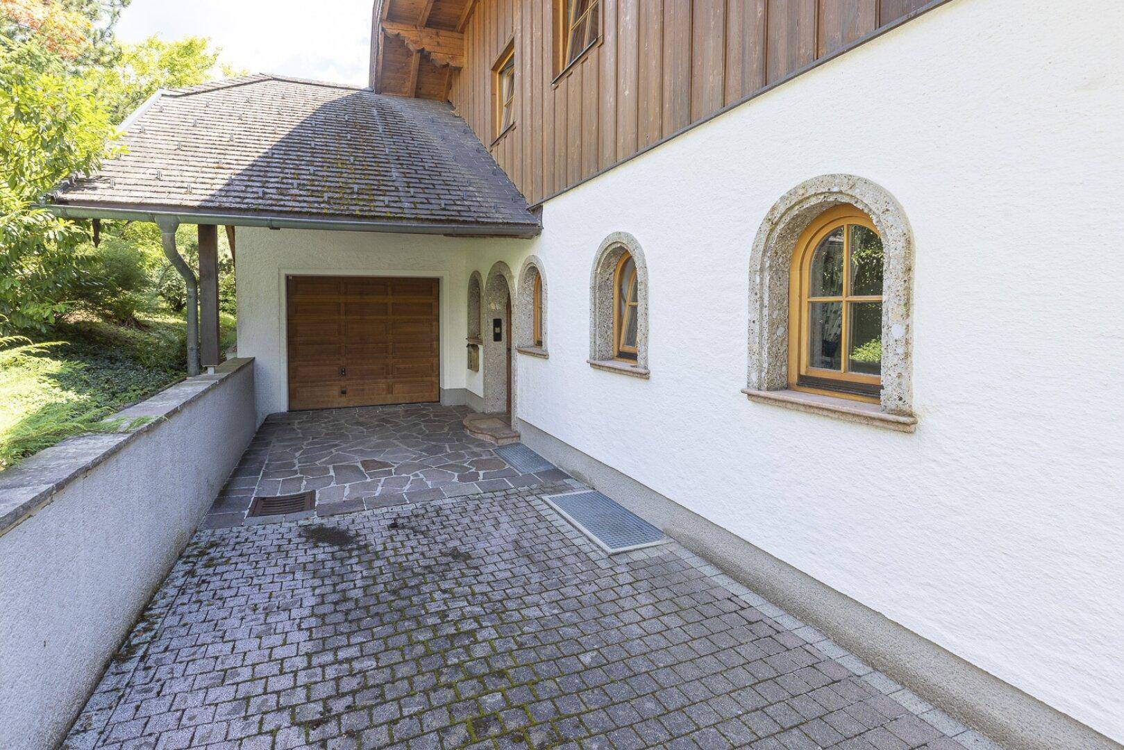 Außenansicht des Hauses mit weißer Fassade, Holztor der Garage und gepflasterter Einfahrt.