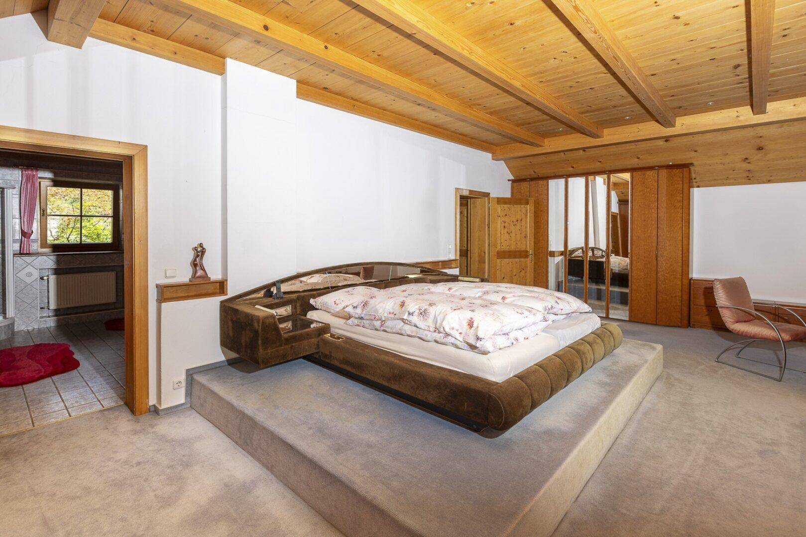 Schlafzimmer mit großem Bett, Teppichboden und Holzbalkendecke, mit Zugang zum Bad.