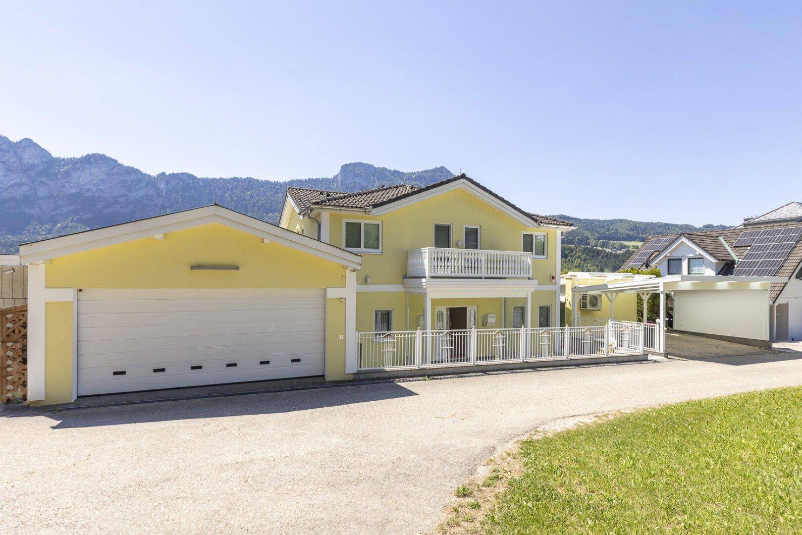 Gesamtansicht des gelben Hauses mit großer Garage und Carport, eingebettet in eine grüne Landschaft.