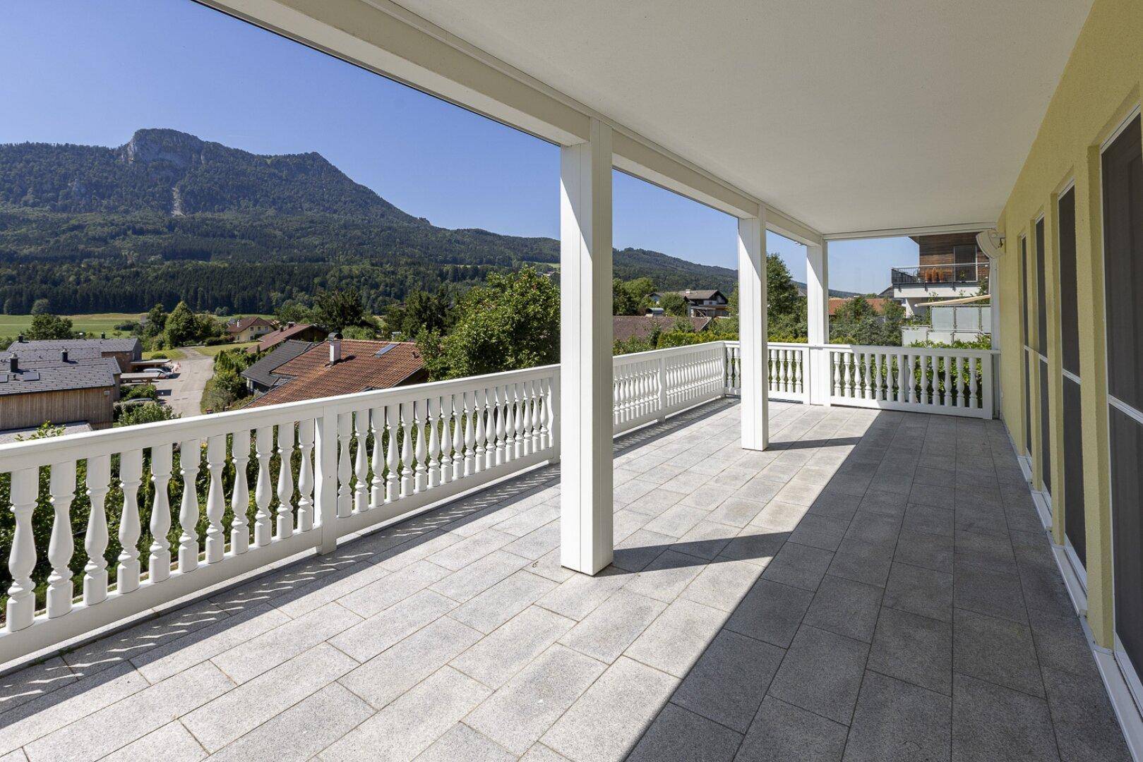 Große überdachte Terrasse mit Fliesenboden und weißem Geländer, bietet einen weiten Blick auf die Berge.