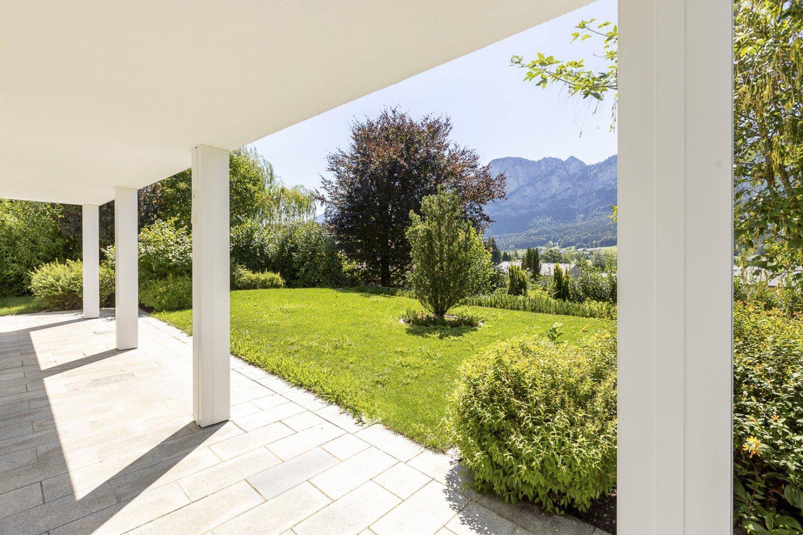 Überdachte Terrasse mit gepflastertem Boden und weitem Blick auf den gepflegten Garten und die Berge.