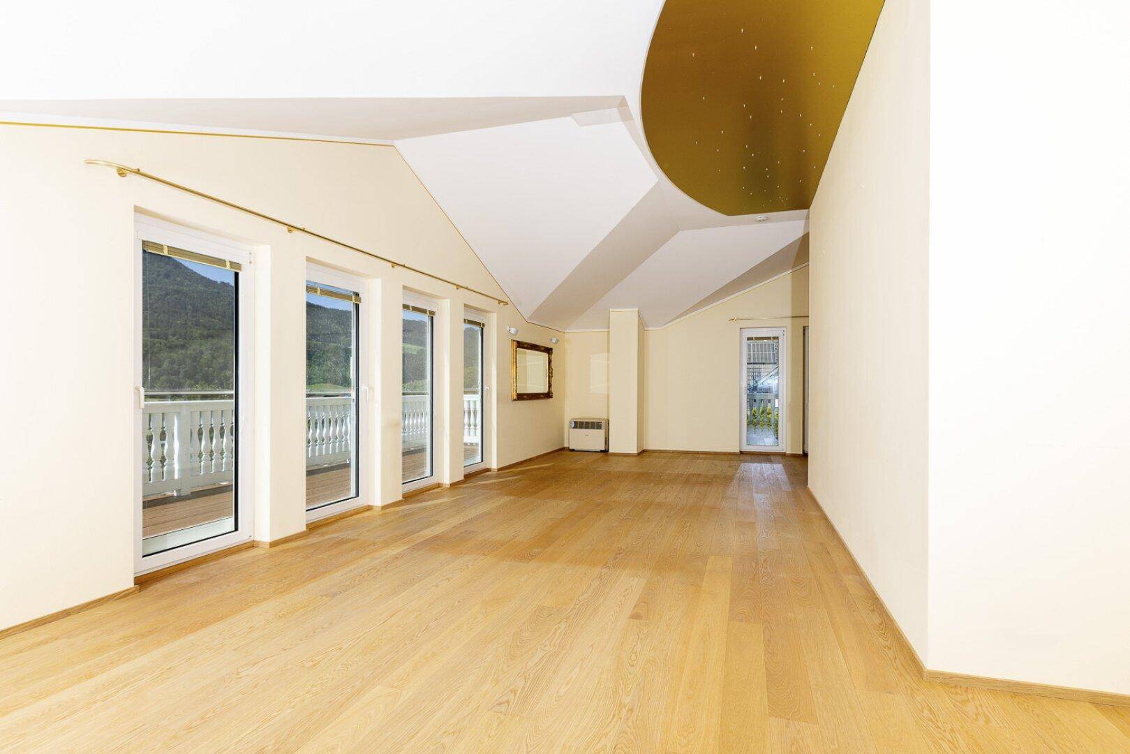 Heller, leerer Raum mit Holzboden, großen Fenstern und Zugang zu einem Balkon mit Bergblick.