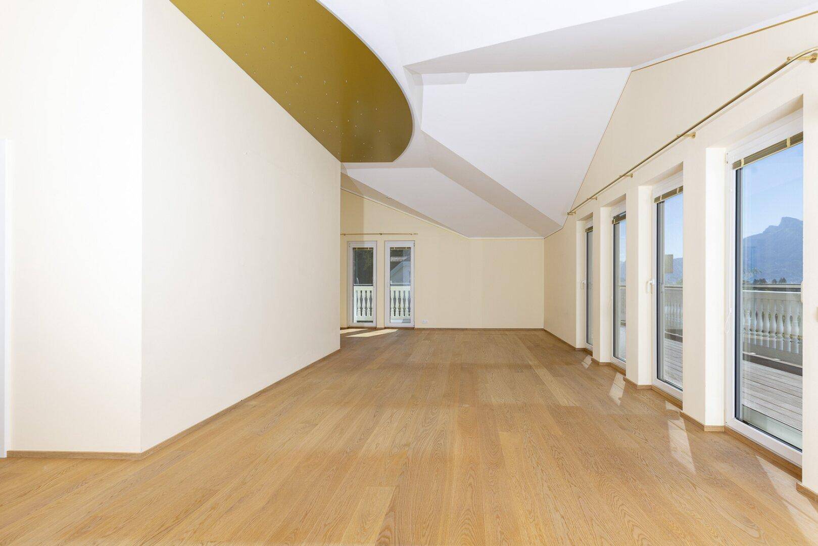 Großzügiger, heller Raum mit Holzboden und mehreren Fenstertüren, die viel Tageslicht hereinlassen.