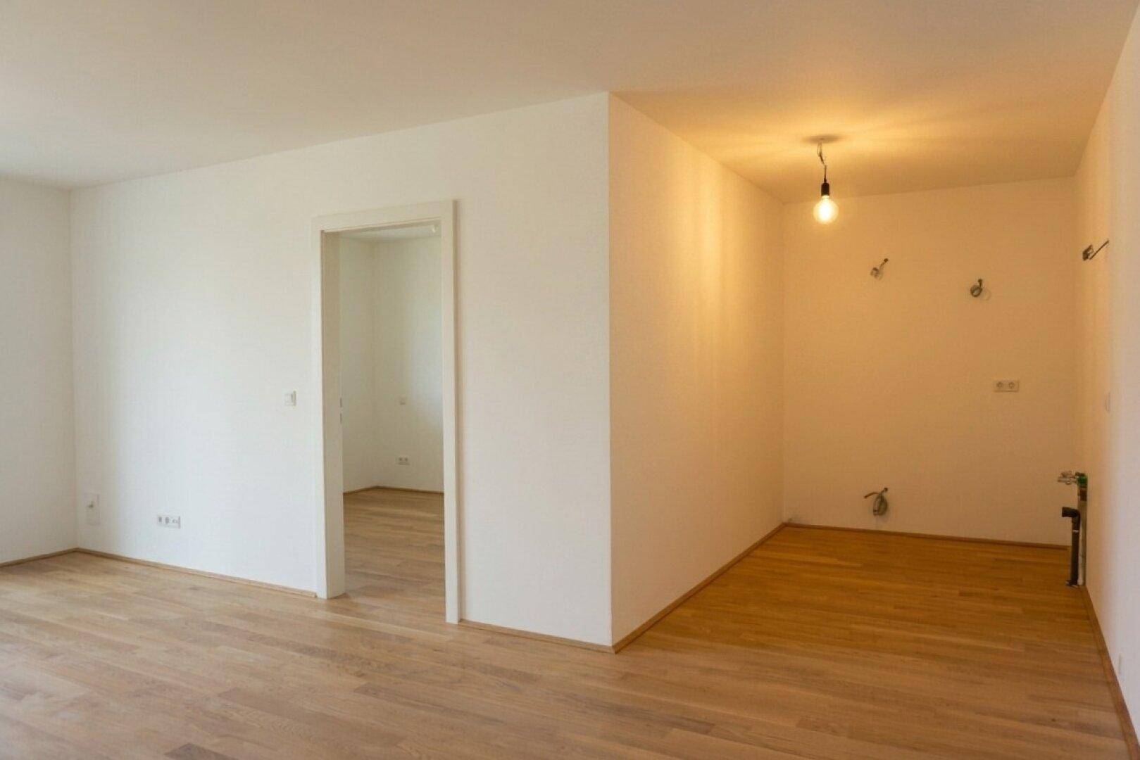 Leerer Küchenbereich mit hellem Holzfußboden und vorbereiteten Anschlüssen für eine Einbauküche.