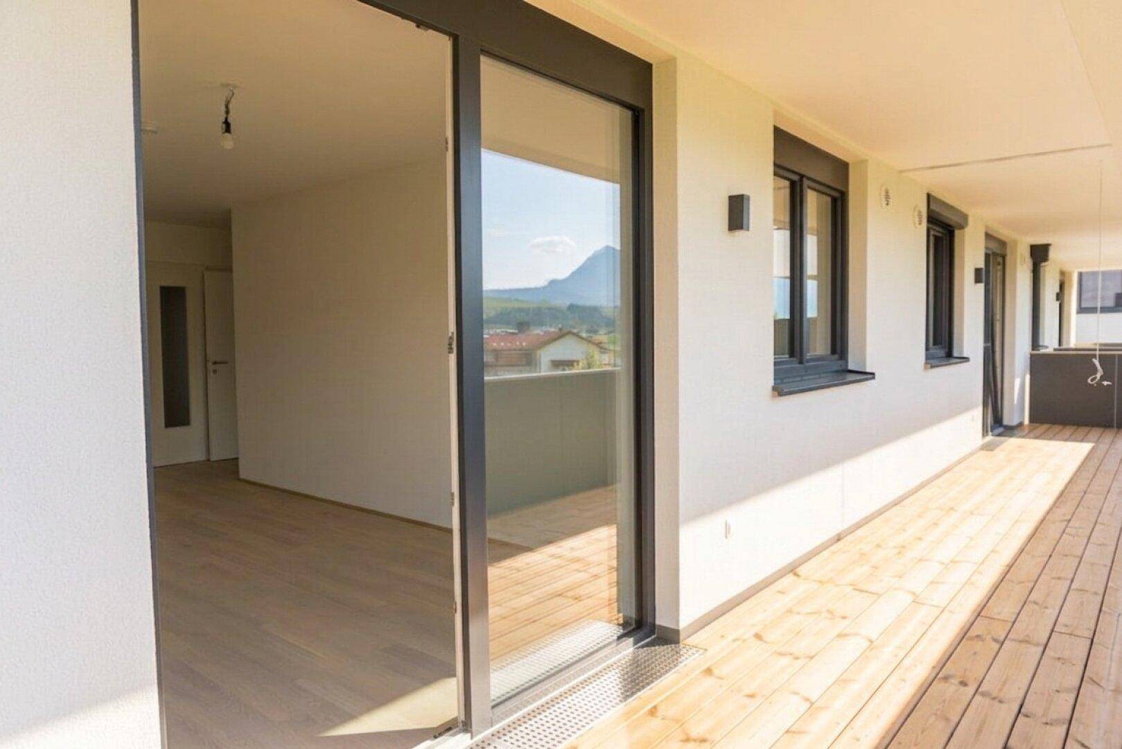 Langer Balkon mit Holzboden und Schiebetür zum Innenbereich, mit Bergblick im Hintergrund.