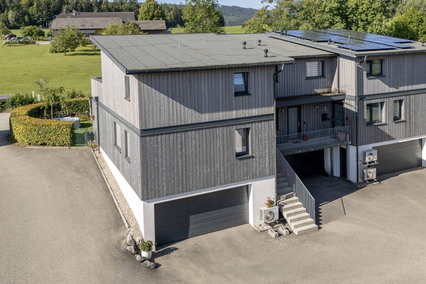 Modernes Haus mit Holzfassade, Flachdach und integrierten Solarpaneelen in ländlicher Umgebung.
