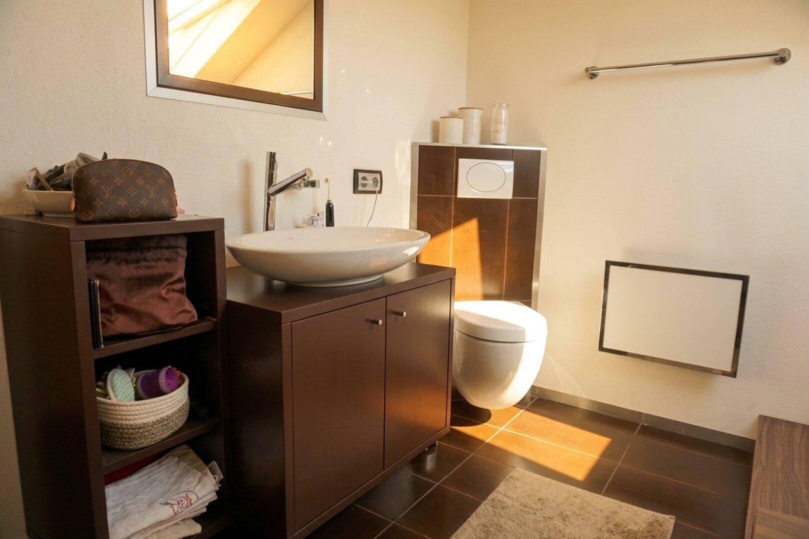 Badezimmer mit Waschtisch, Toilette und dunklen Fliesen, gut beleuchtet.