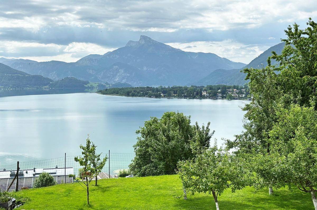 Malerischer Blick über einen See und Berge von einem grünen Garten mit jungen Bäumen.