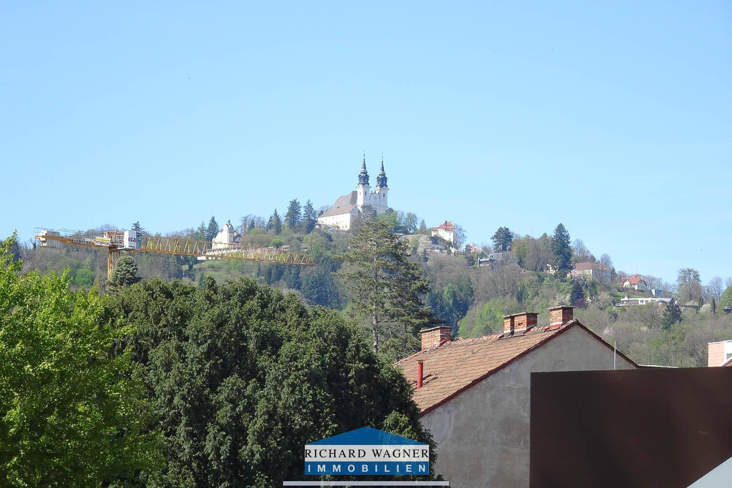Panoramablick auf eine historische Kirche auf einem bewaldeten Hügel mit grünen Bäumen im Vordergrund.