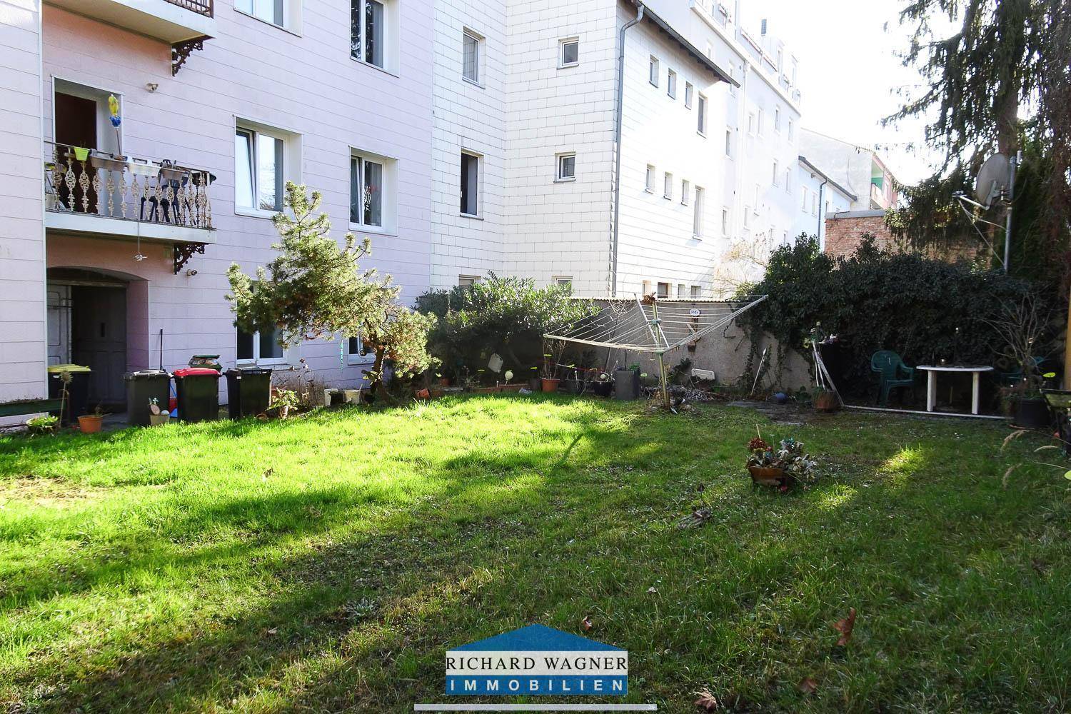 Grüner Hinterhofgarten mit Rasenfläche, Sträuchern und einem kleinen Gewächshaus hinter dem Gebäude.