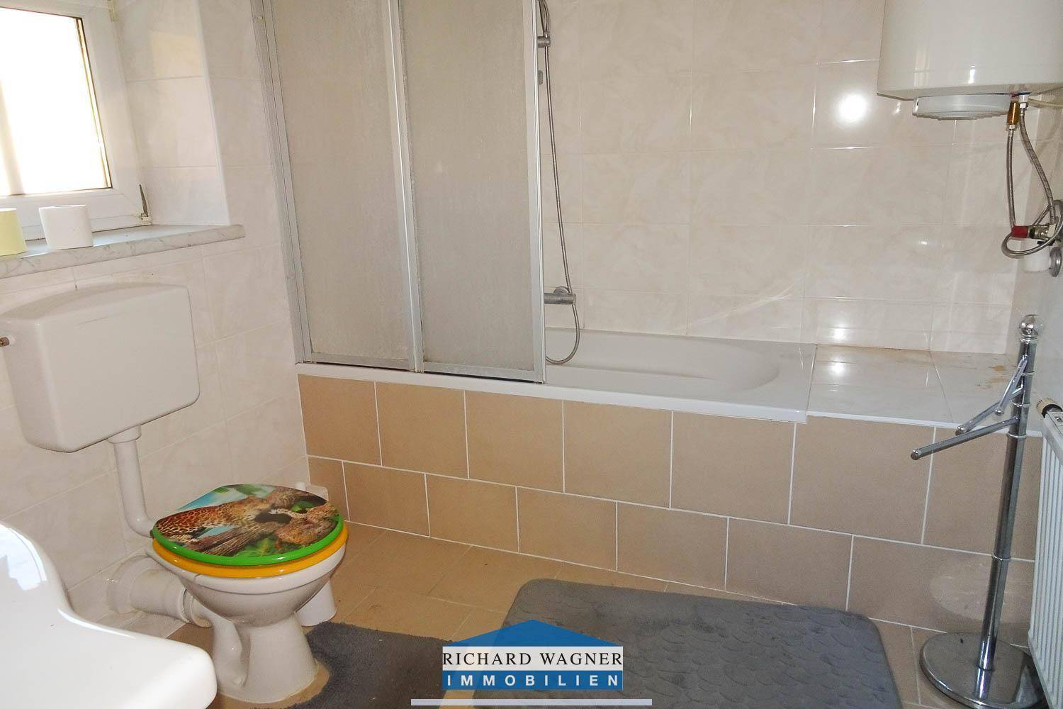 Badezimmer mit Badewanne, Duschabtrennung, WC und Fenster, komplett gefliest in hellen Farbtönen.