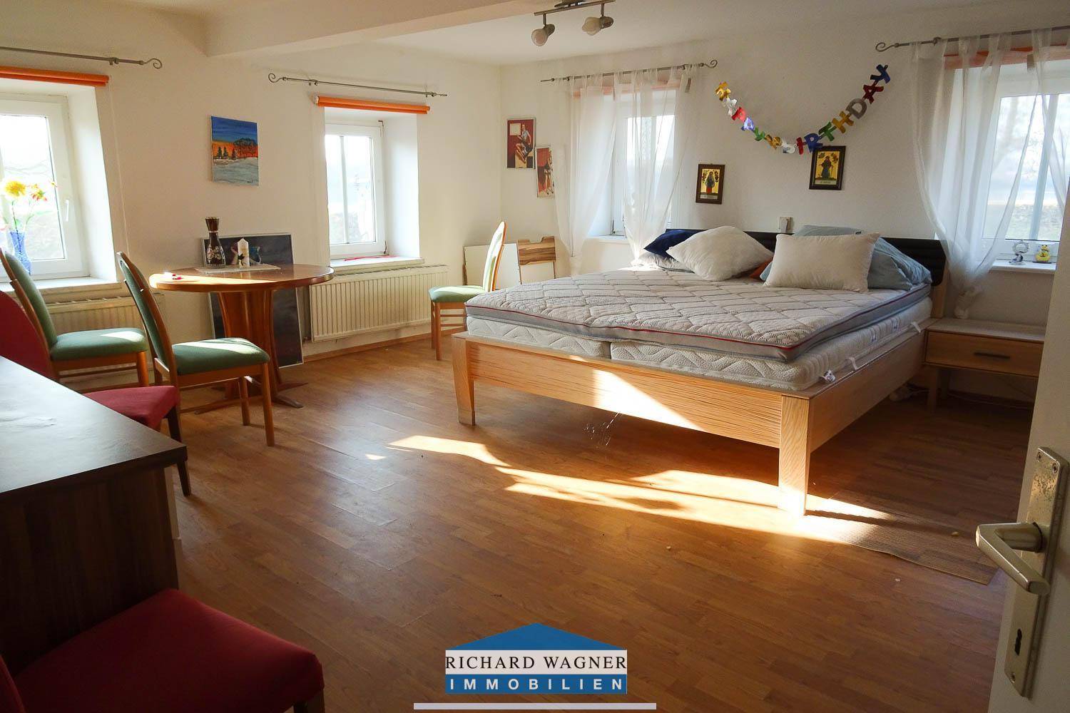 Helles Schlafzimmer mit Holzboden, Doppelbett und mehreren Fenstern, die viel Tageslicht hereinlassen.