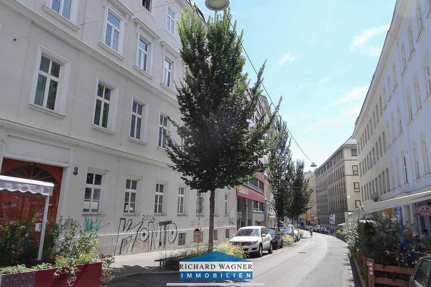 Belebte Stadtstraße mit Bäumen, parkenden Autos und Gebäuden entlang des Gehwegs unter blauem Himmel.