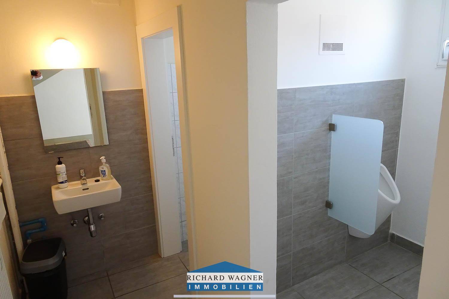 Toilettenraum mit Urinal, Waschbecken und grauen Fliesen an den Wänden.