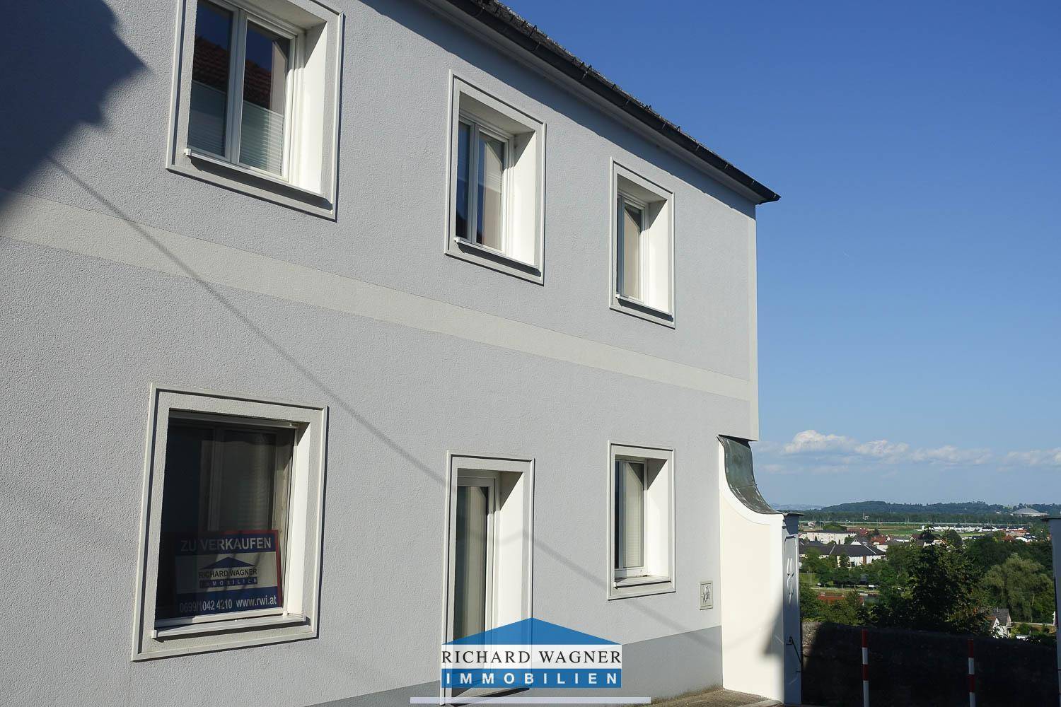 Seitenansicht des Gebäudes mit hellblauer Fassade und einem "Zu verkaufen"-Schild im Fenster.