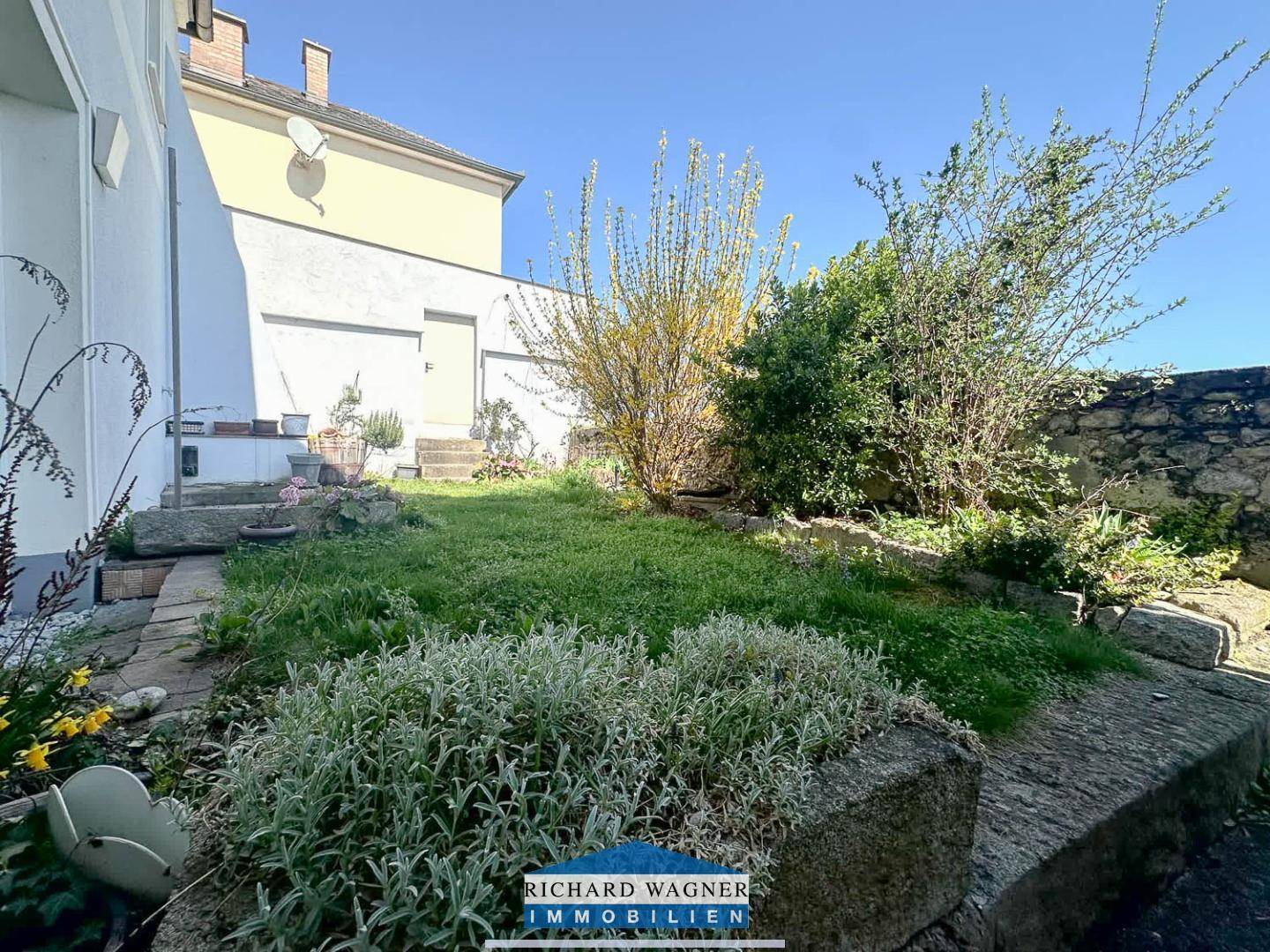 Sonnige Gartenecke mit grüner Rasenfläche, Steinbegrenzungen und blühenden Pflanzen.