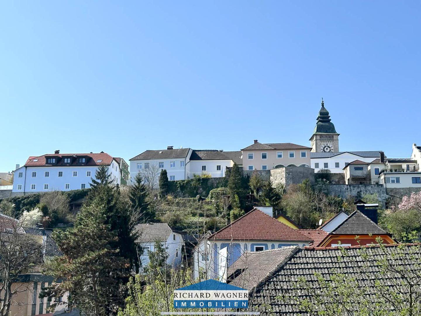 Blick auf eine malerische Stadtansicht mit historischen Gebäuden, einem Kirchturm und grünen Hügeln unter blauem Himmel.