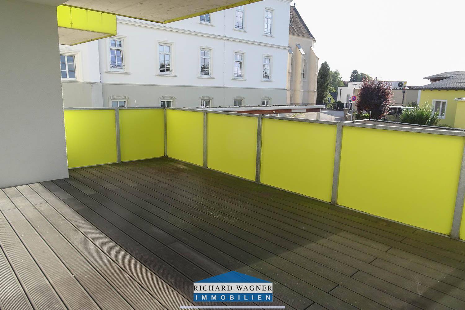 Heller Balkon mit Holzboden und gelber Brüstung, bietet einen Blick auf ein klassisches Gebäude.
