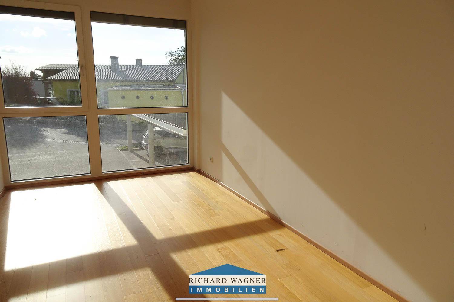 Helles Zimmer mit Holzboden und großen Fenstern, bietet einen Blick auf den Carport und die Umgebung.