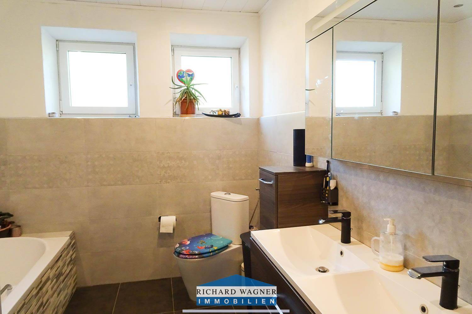 Badezimmer mit Badewanne, WC, Einzelwaschbecken und zwei Fenstern.