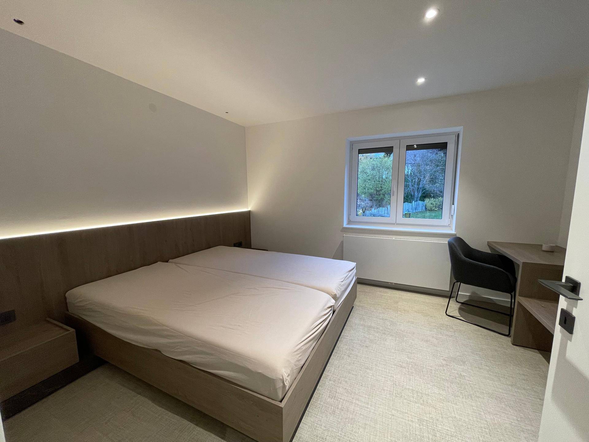 Helles Schlafzimmer mit Doppelbett, Schreibtisch und Blick ins Grüne durch das Fenster.