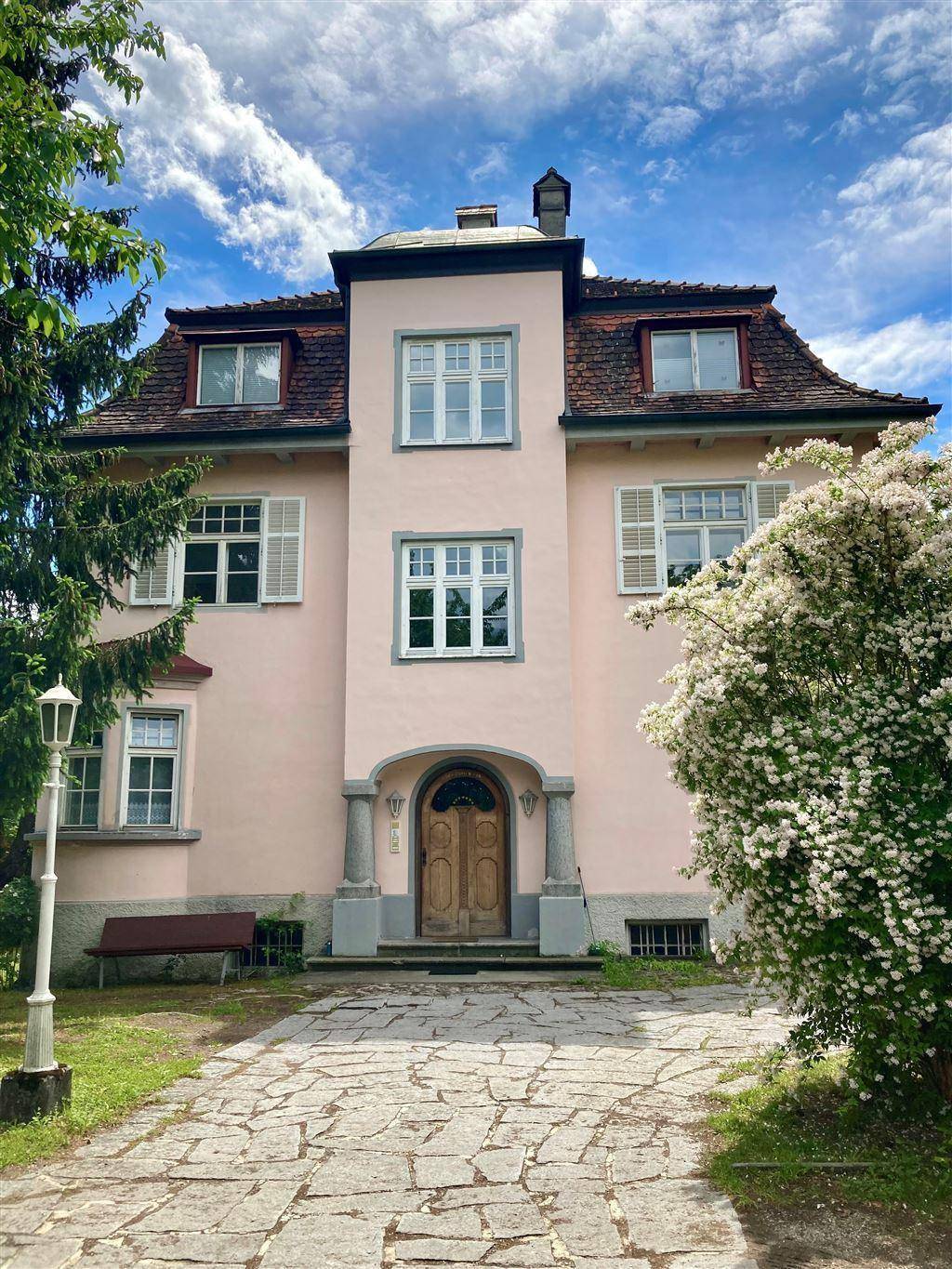 Vorderansicht der historischen Villa mit gepflastertem Zugang und blühenden Sträuchern im Garten.