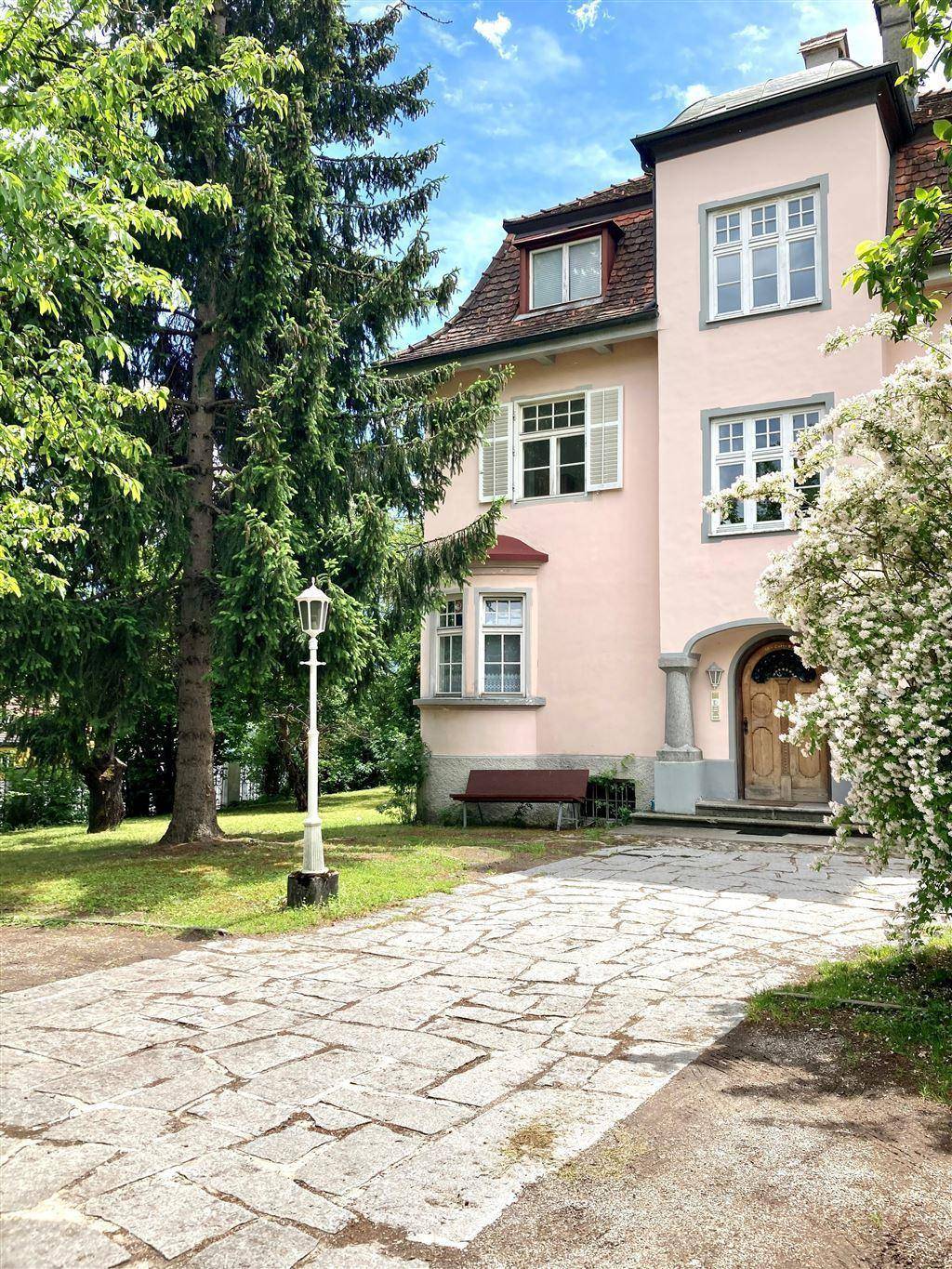 Seitenansicht der historischen Villa mit hohen Bäumen und einem gepflasterten Weg zum Eingangsbereich.