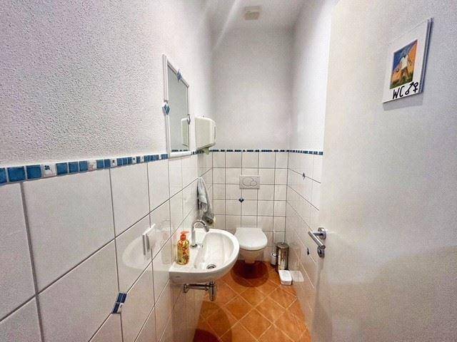 Kompaktes WC mit weißen Wandfliesen, Terrakotta-Boden und einem kleinen Waschbecken und Spiegel.
