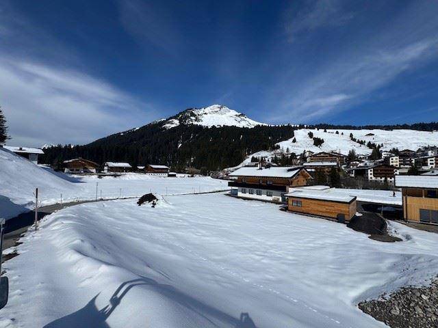 Winterlandschaft mit schneebedeckten Wiesen und Häusern vor einer Bergkulisse.