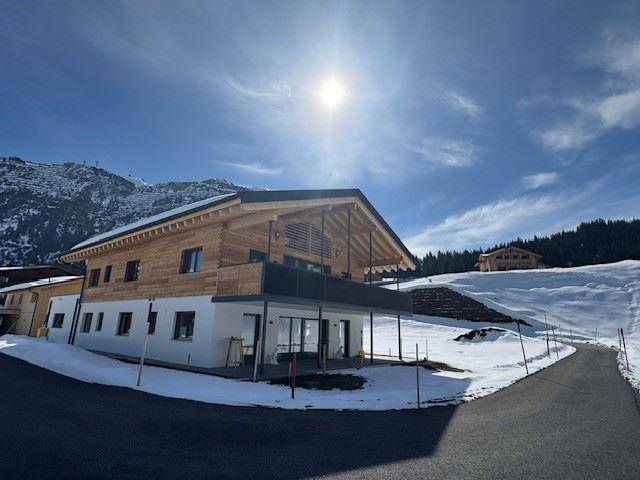Modernes Holzhaus mit Balkon in sonniger Winterlandschaft, umgeben von Schnee und Bergen.