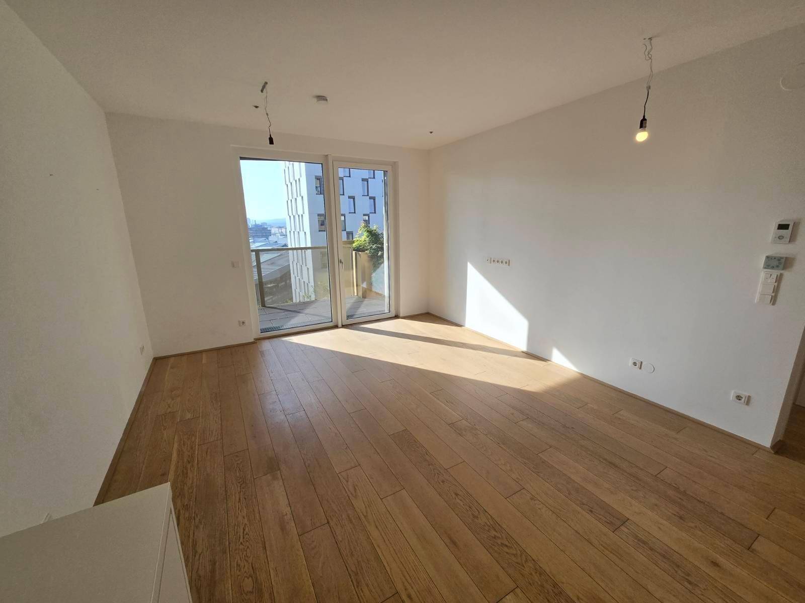 Lichtdurchfluteter Wohnbereich mit Holzboden und großen Fenstern, die Zugang zum Balkon ermöglichen.