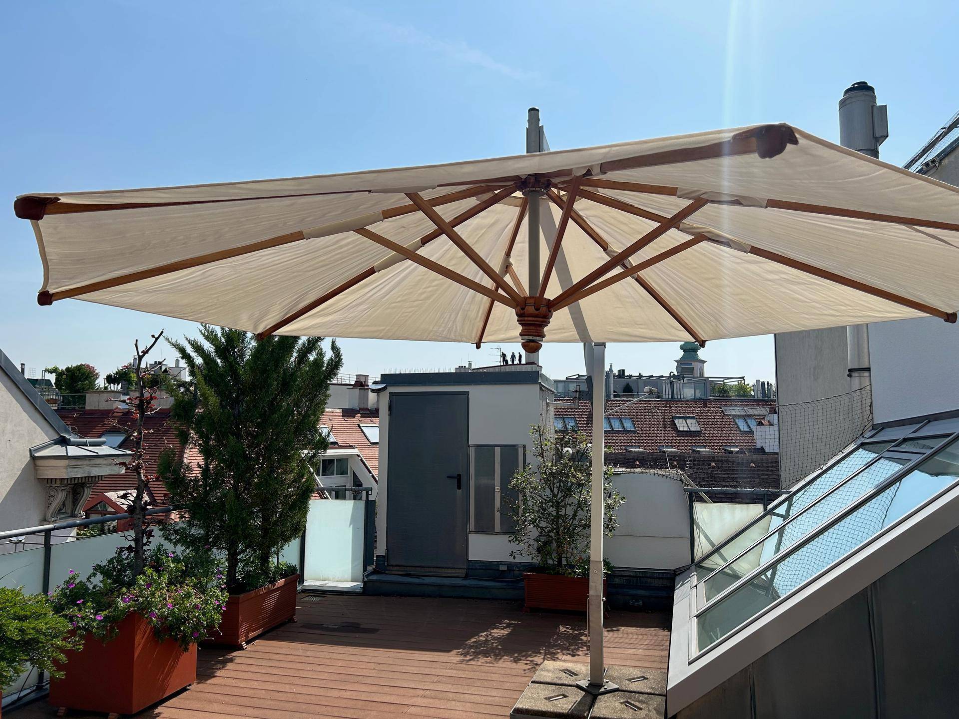 Sonniger Dachgarten mit Holzdeck, Pflanzen und großem Sonnenschirm unter blauem Himmel.