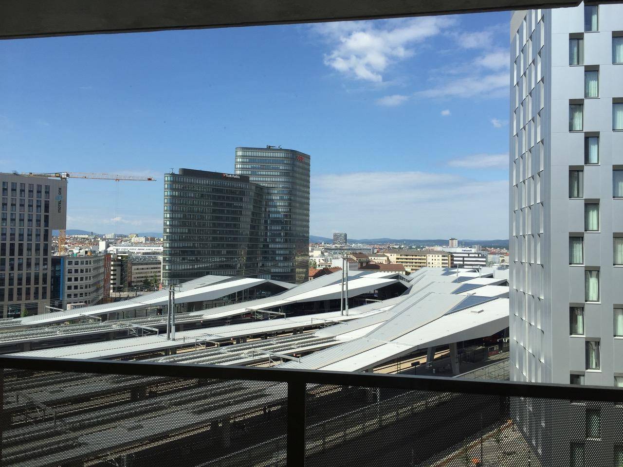 Panoramablick von einem Balkon auf eine moderne Stadtlandschaft mit Bahnhof und Hochhäusern unter blauem Himmel.