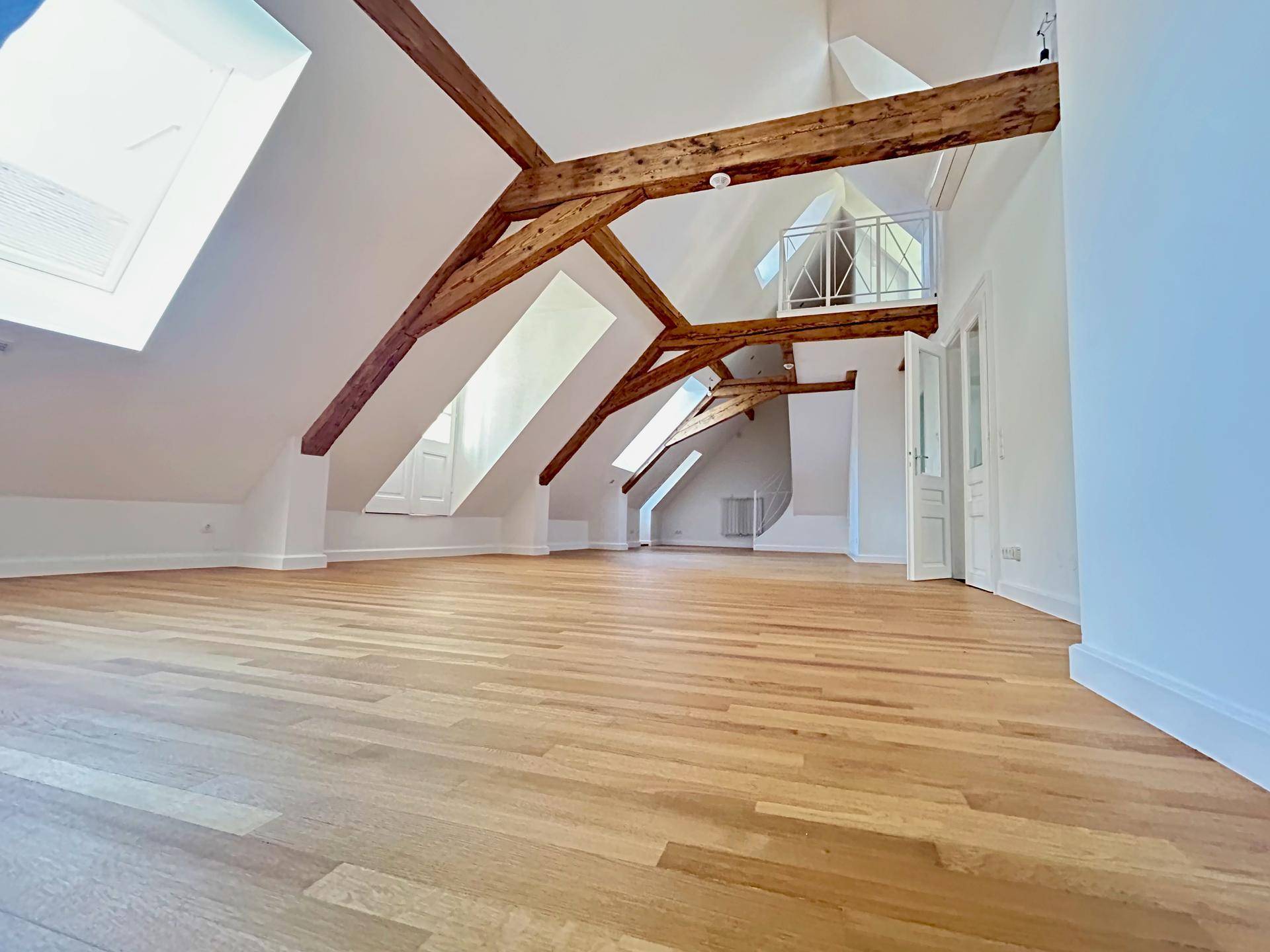 Lichtdurchfluteter Dachgeschossraum mit Holzbalken, Parkettboden und mehreren Dachfenstern.