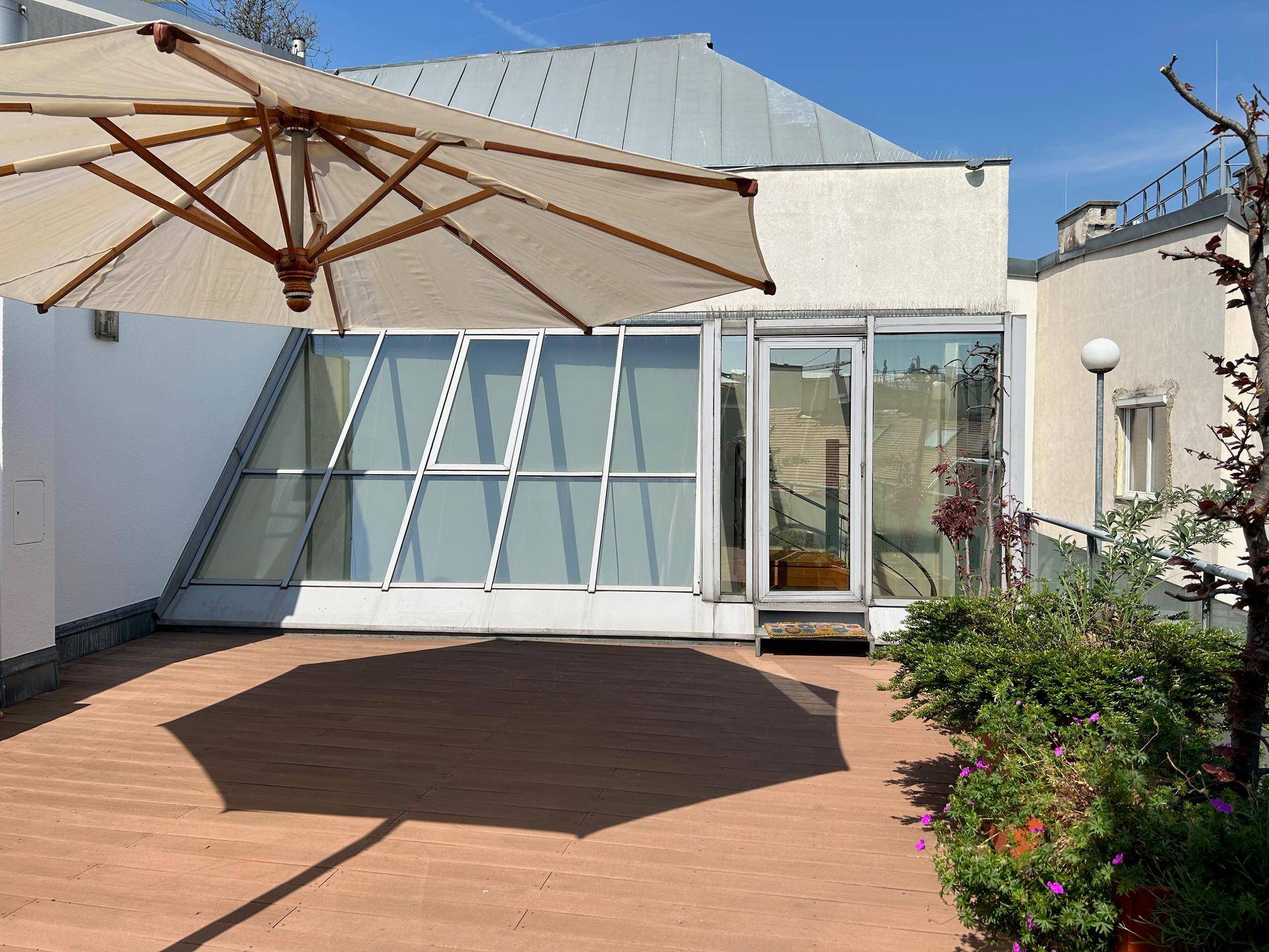 Weitläufige Dachterrasse mit Holzdeck, Pflanzen und Sonnenschirm, umgeben von moderner Architektur.