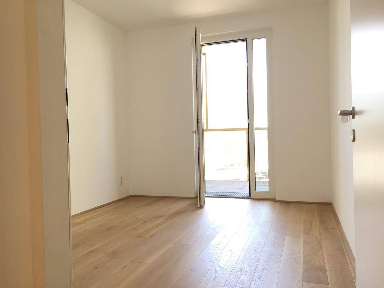 Helles Schlafzimmer mit Holzboden und offener Balkontür, die viel Tageslicht hereinlässt.