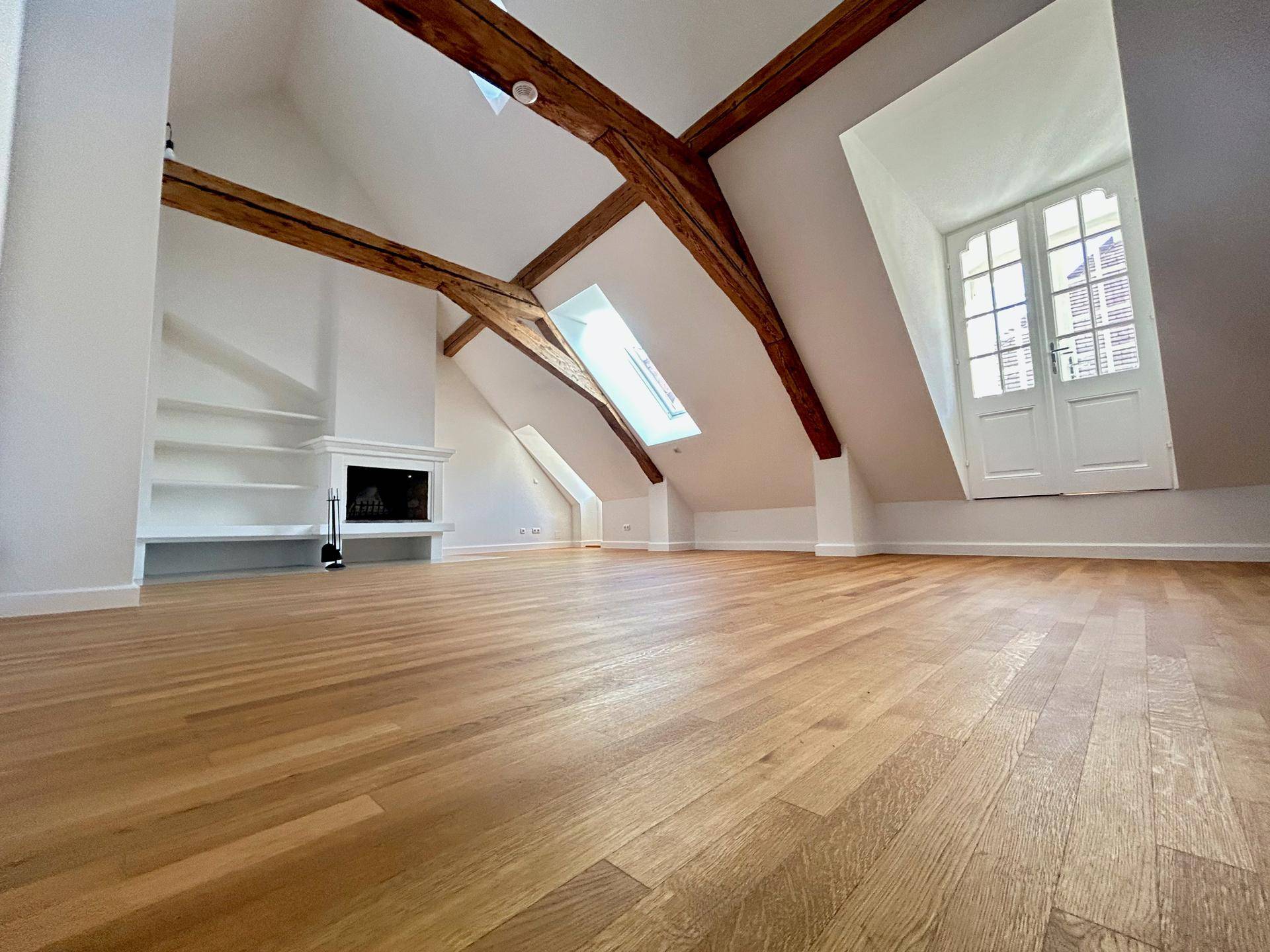Helles Dachgeschosszimmer mit Holzbalken, Parkettboden, Kamin und Dachfenstern.