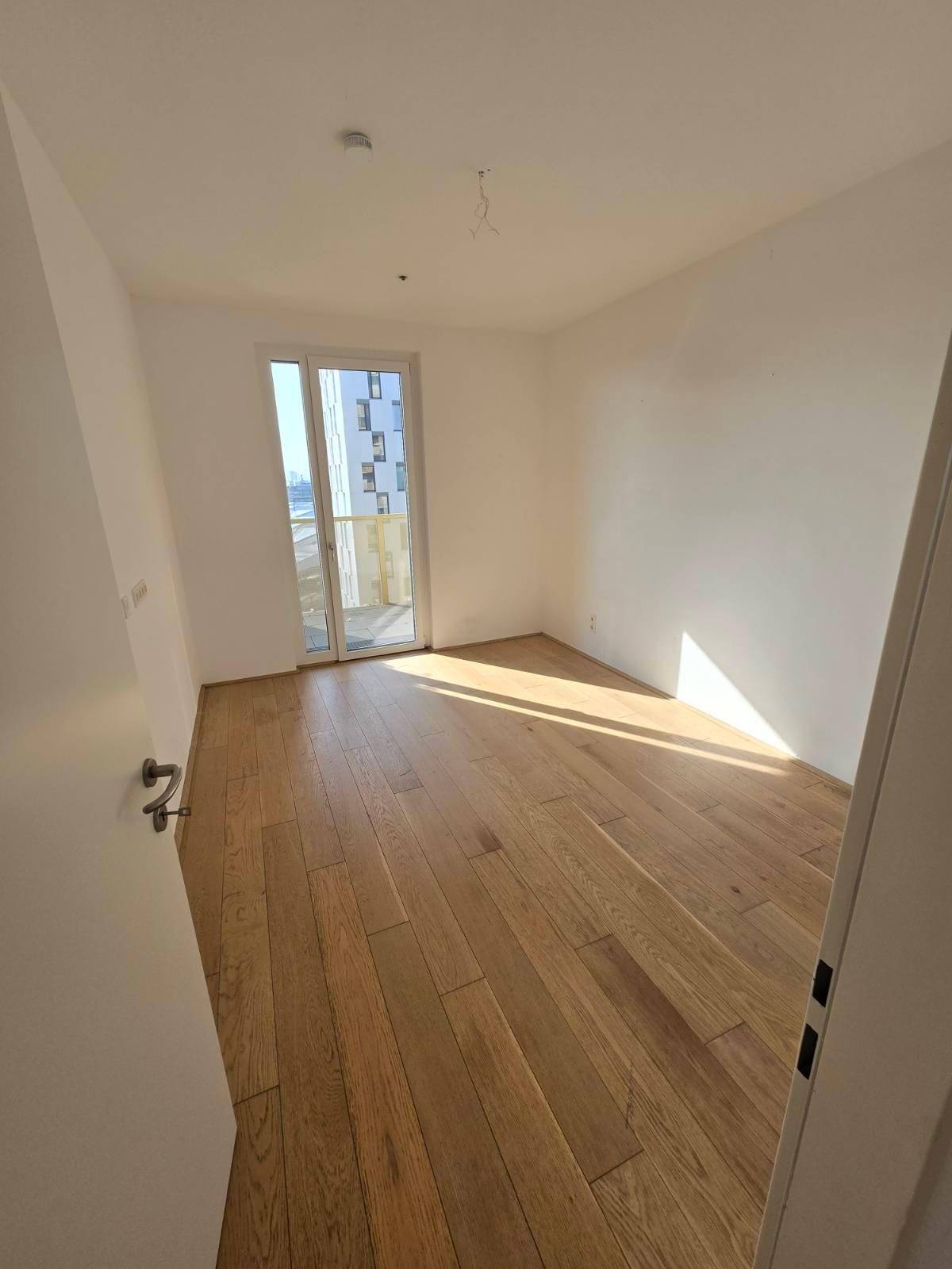 Helles Schlafzimmer mit Holzboden und großem Fenster, das Zugang zum Balkon bietet.